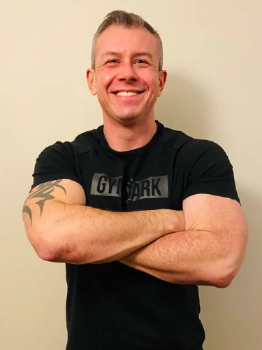 A portrait of Frank, a fitness instructor at East Kilbride Taekwon-Do & Fitness Academy. He is smiling warmly with his arms crossed, wearing a black Gymshark athletic t-shirt that shows a tribal-style tattoo on his right arm. He is standing against a neutral, light-colored background.