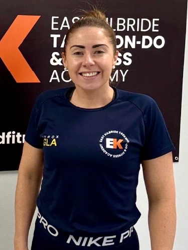 A portrait of Stacey Cummings, a fitness instructor at the academy. She is smiling and wearing a dark blue athletic t-shirt with the "EK" East Kilbride Taekwon-Do & Fitness Academy logo on the chest. She stands in front of a black and orange academy banner that reads "East Kilbride Taekwon-Do & Fitness Academy.