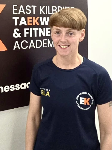 A portrait of Kristen Meechan, a fitness instructor at the academy. She is smiling and wearing a dark blue athletic t-shirt with the "EK" East Kilbride Taekwon-Do & Fitness Academy logo on the chest. She stands in front of a black and orange academy banner that reads "East Kilbride Taekwon-Do & Fitness Academy.