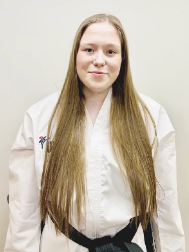 A portrait of Miss Feeney, a Taekwon-Do instructor, standing in front of a neutral white wall. She is wearing a traditional white Taekwon-Do dobok with black trim and a black belt. The uniform features the UKTC logo on the right chest and the International Taekwon-Do Federation (ITF) patch on the left.