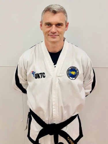 A portrait of Mr. Stewart, a Taekwon-Do instructor, standing in front of a neutral white wall. He is wearing a traditional white Taekwon-Do dobok with black trim and a black belt. The uniform features the UKTC logo on the right chest and the International Taekwon-Do Federation (ITF) patch on the left. He is smiling slightly with his hands held behind his back in a formal stance.